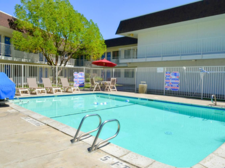 A swimming pool area with lounge chairs, umbrellas, and a surrounding fence in a sunny courtyard setting.