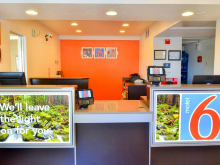 This image shows the reception area of a Motel 6, featuring a front desk with branding signs and bright lighting.