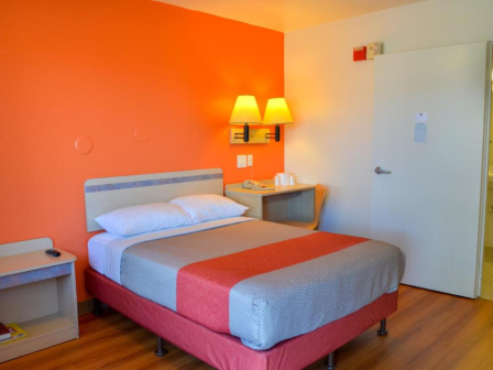 A hotel room with a bed, nightstand, and a bright orange accent wall. There's a door leading to the bathroom on the right.